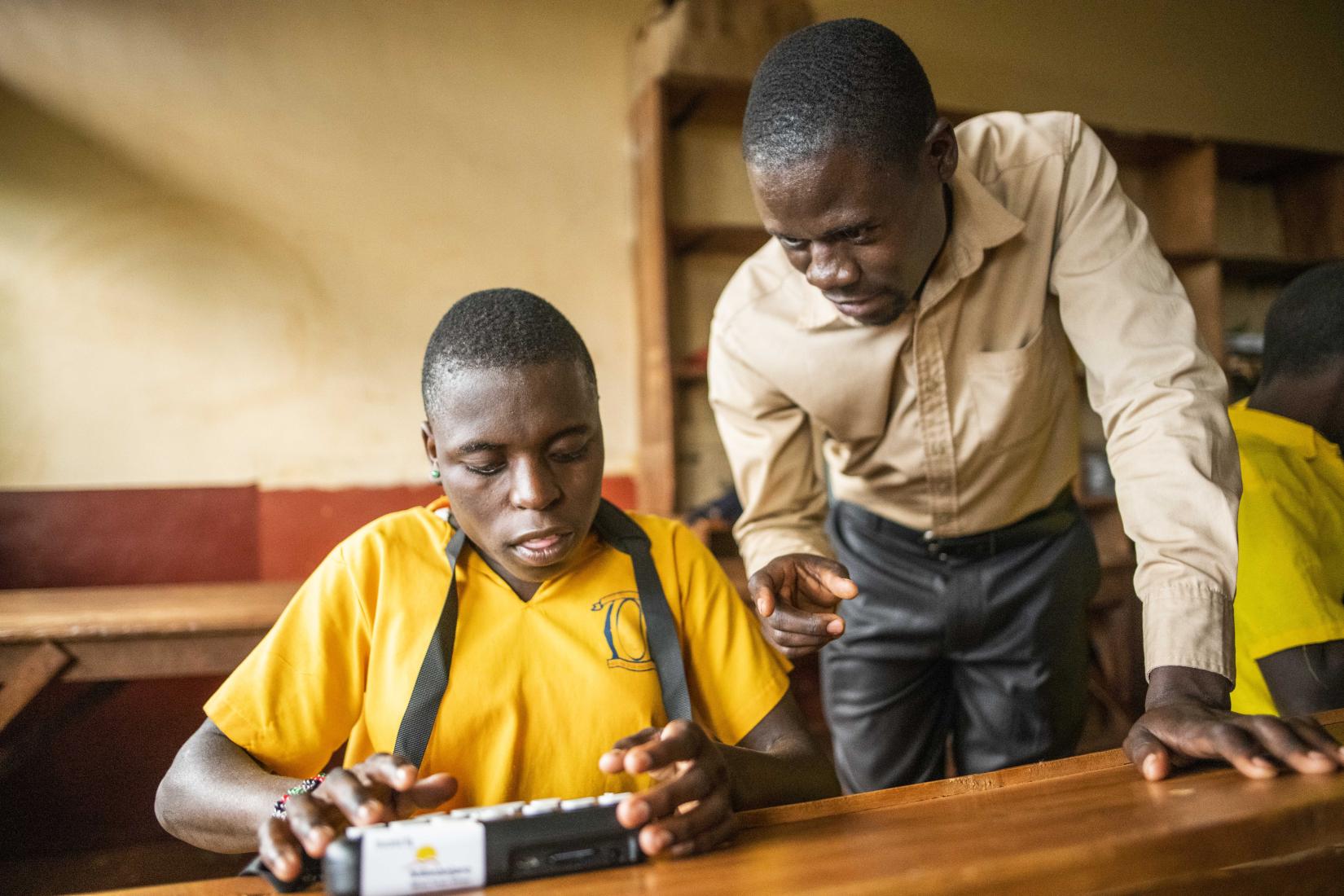 Special needs teacher Tiwaku John from Spire Road Primary school in Jinja assists blind student Sarah Musasizi, 13yrs, on the Orbit braille reader. 